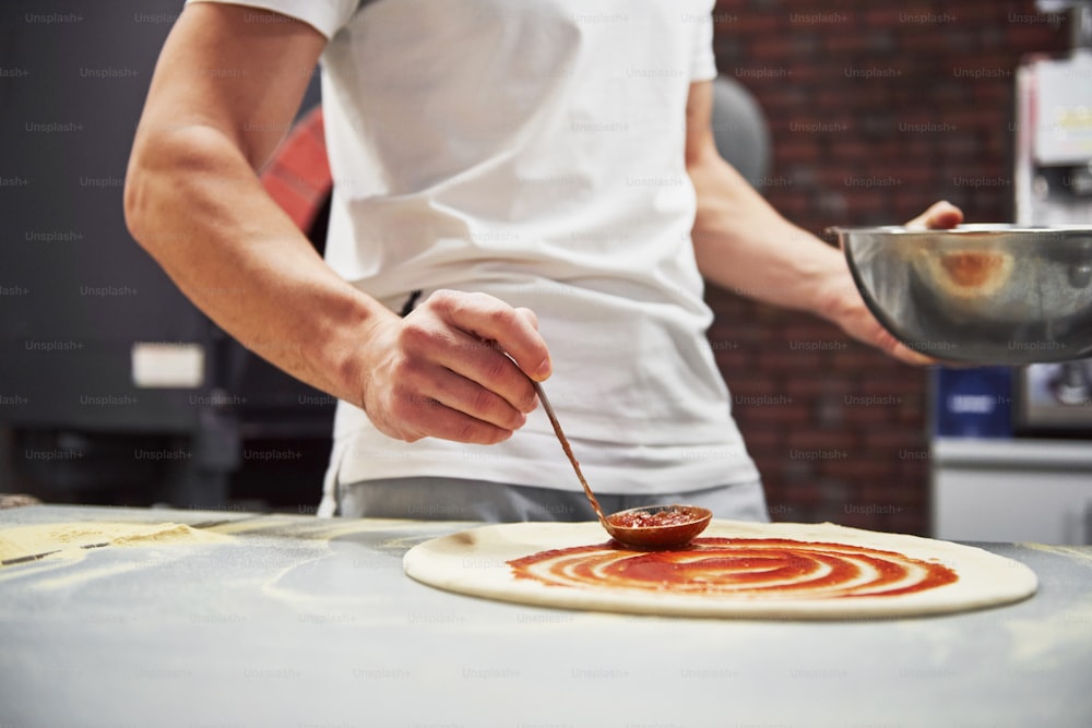 Nahaufnahme des Bäckers, der Sauce legt, um köstliche Pizza für eine Bestellung im Restaurant zu machen.