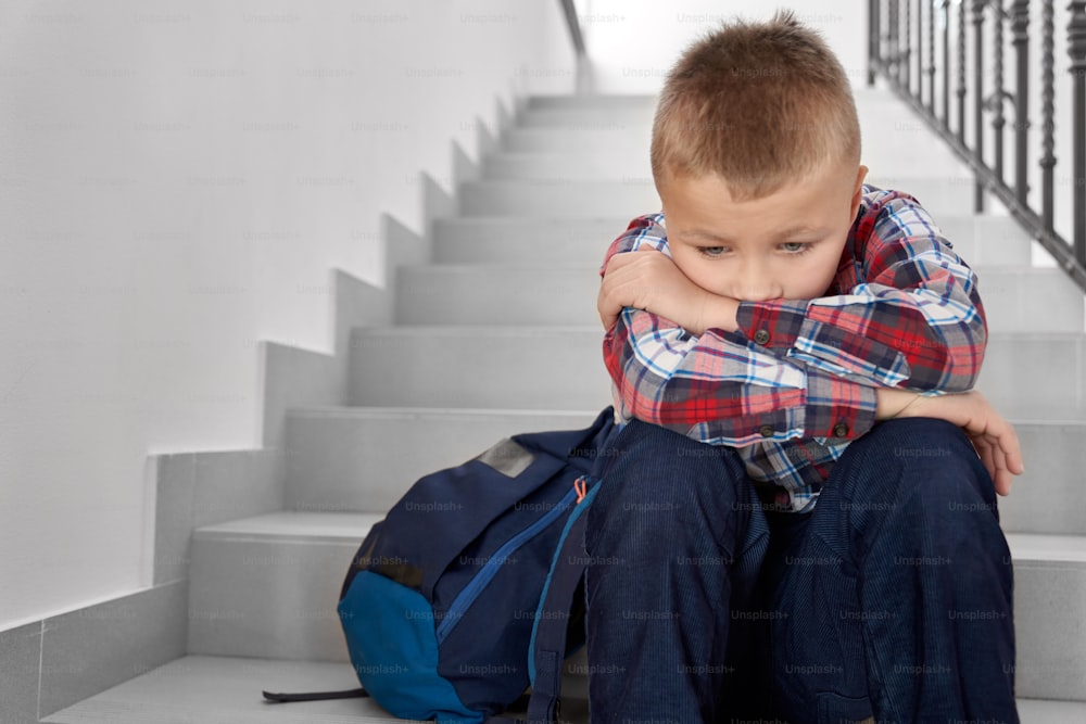 Decepcionado, triste colegial de primaria con mochila sentada en el hueco de la escalera. Un escolar guapo y espesado que vestía una camisa a cuadros y apoyaba la cabeza en las manos.