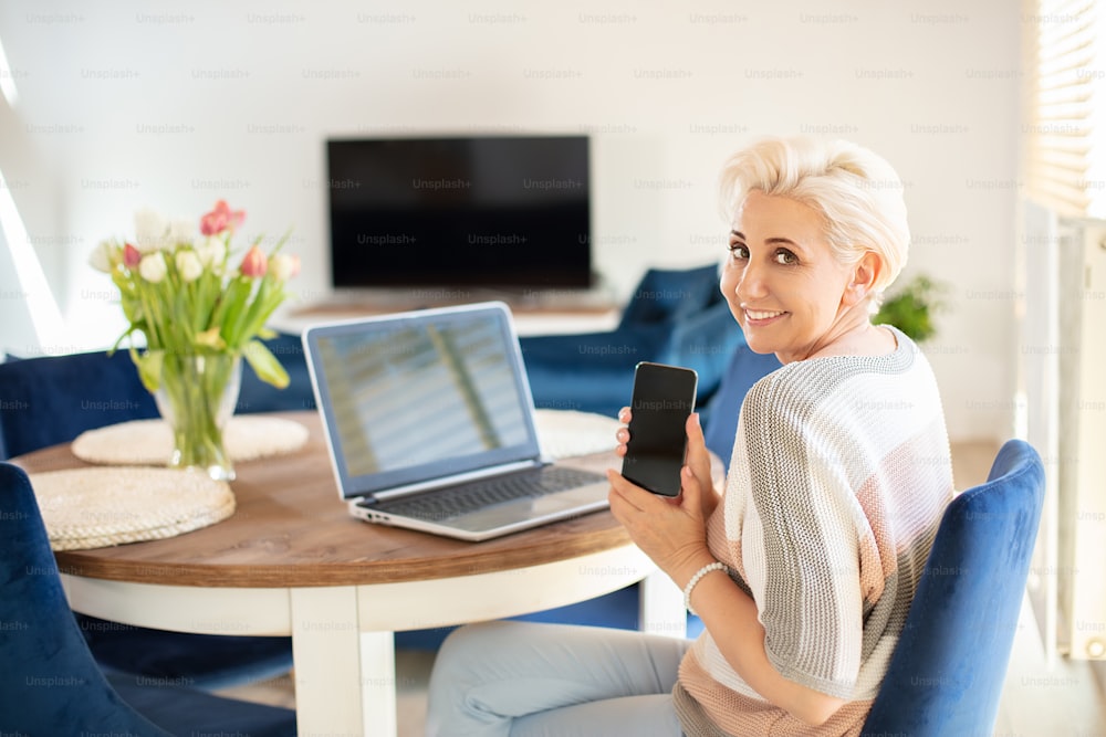 Beautiful blonde woman using laptop and mobile phone , showing empty screen. Happy lady working in home office. Middle age woman smiling to the camera.