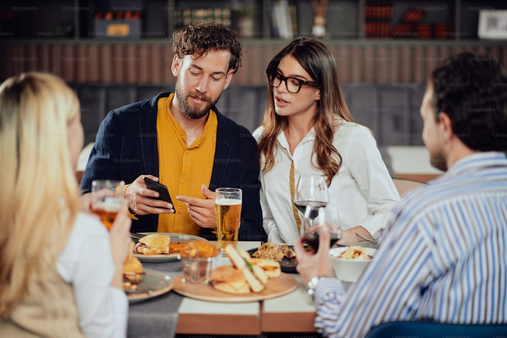 Young attractive Caucasian couple sitting in restaurant and looking at smart phone. In foreground their friends sitting adn drinking.