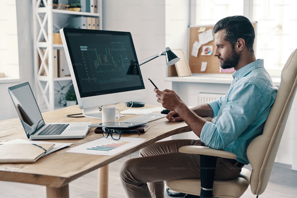 Junger moderner Geschäftsmann mit Smartphone, während er im Büro sitzt