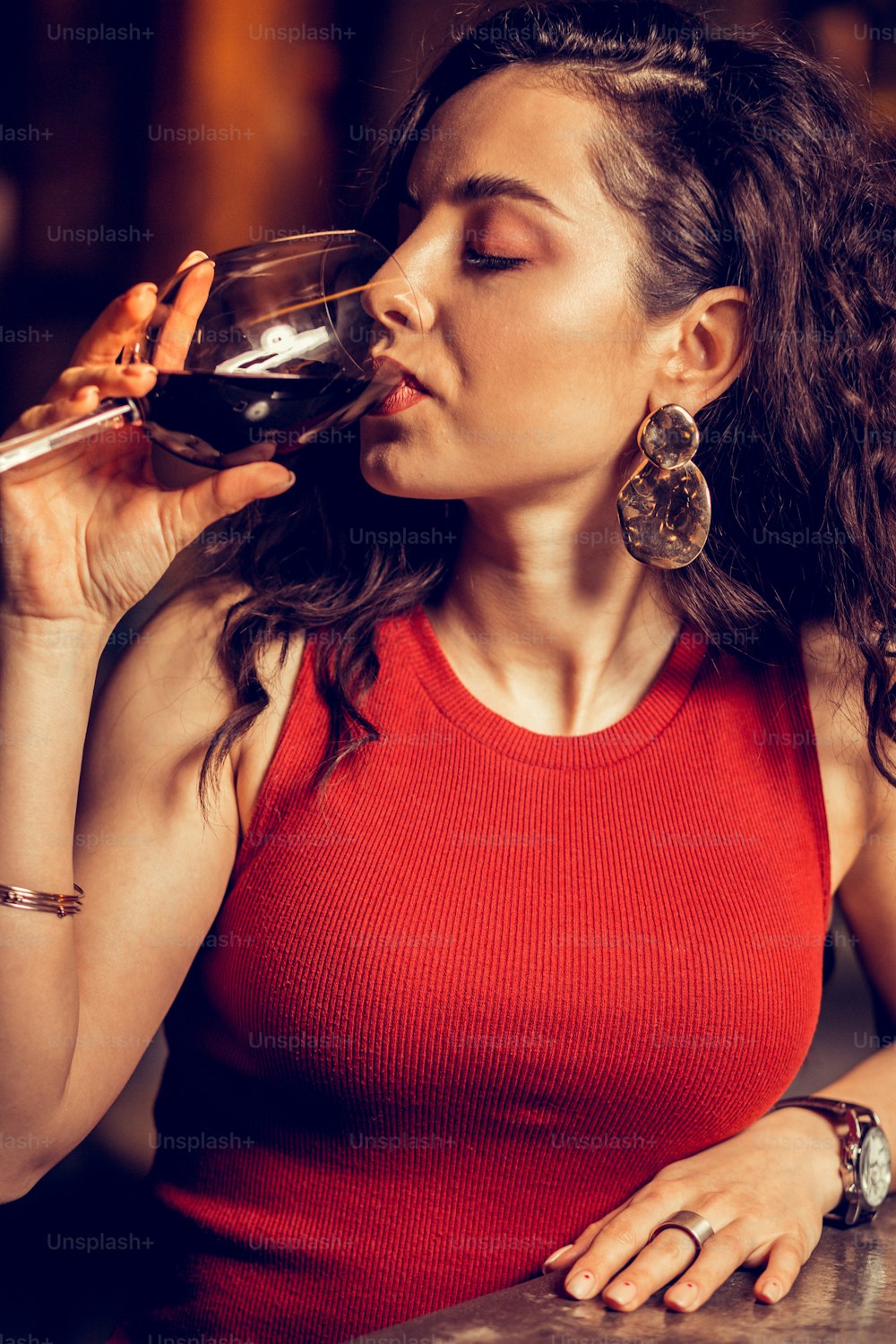 Wein trinken. Nahaufnahme einer lockigen Frau in rotem Kleid, die Rotwein trinkt, während sie allein in der Bar sitzt