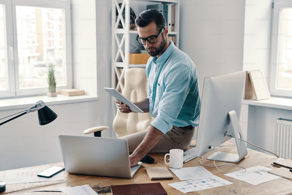 Junger moderner Geschäftsmann mit digitalem Tablet und Laptop während der Arbeit im Büro