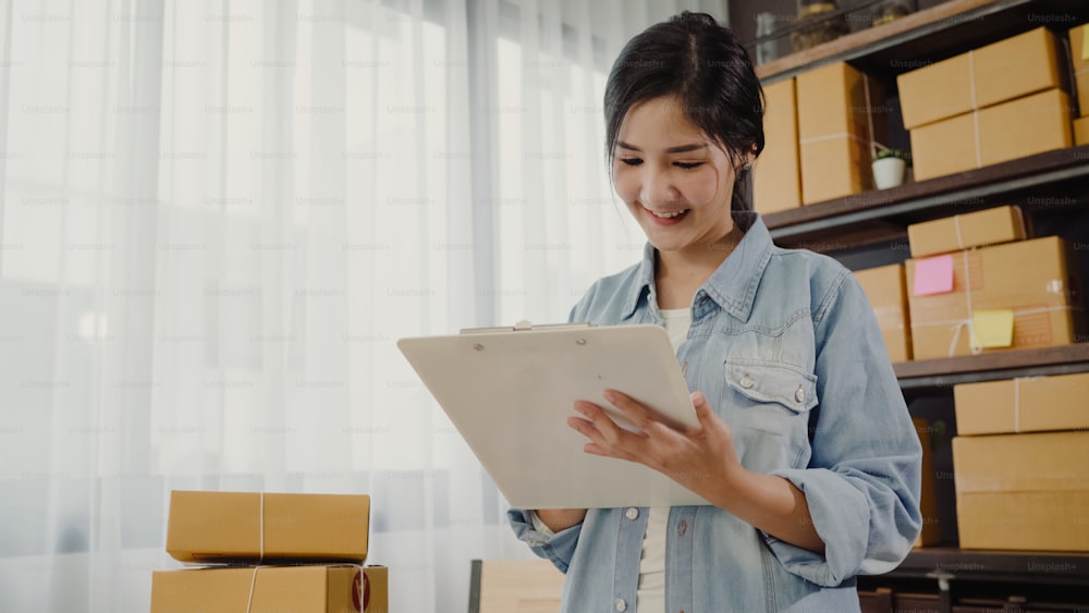 Beautiful smart Asian young entrepreneur business woman owner of SME checking product on stock and write on clipboard working at home. Small business owner at home office concept.