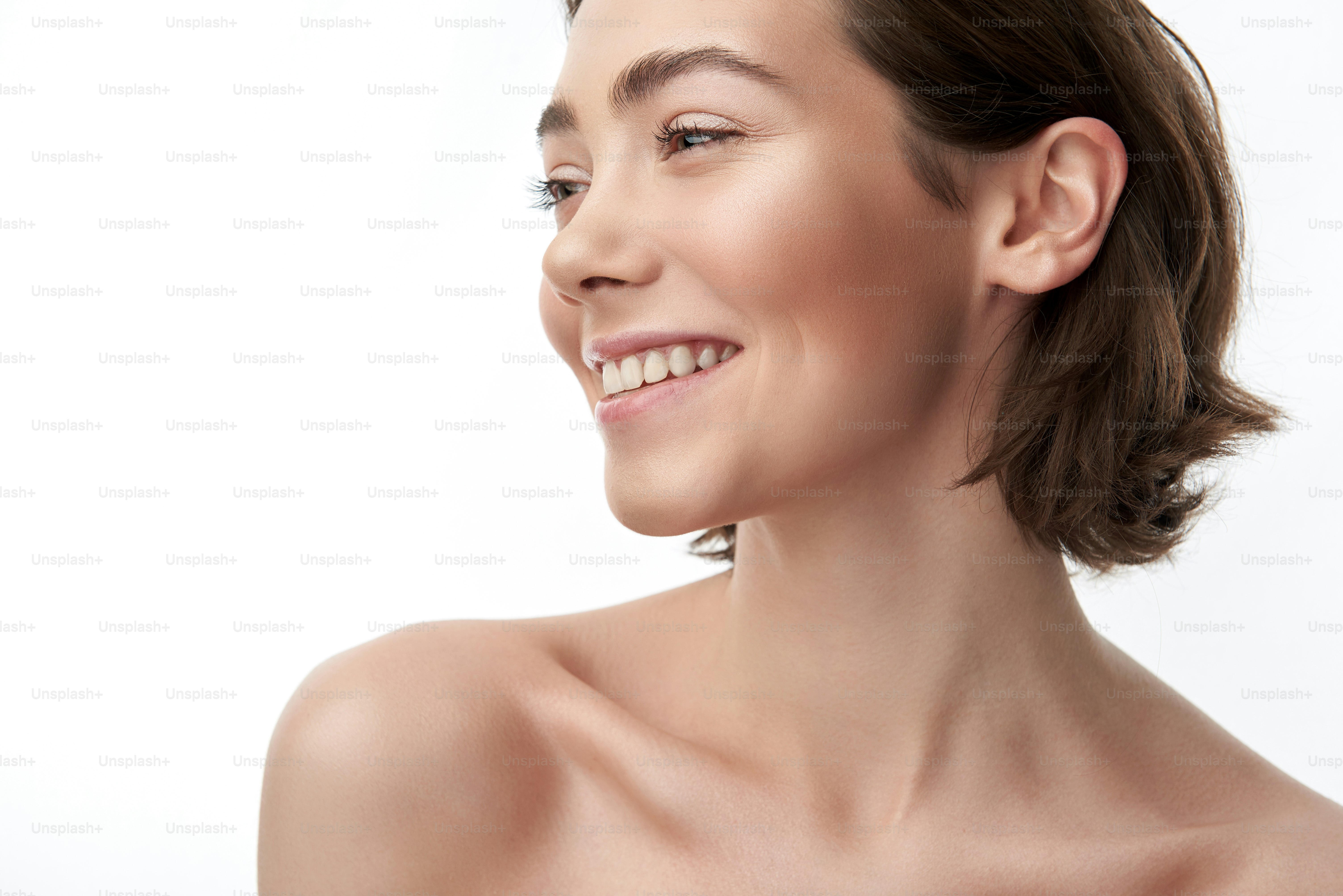 Nahaufnahme Porträt einer jungen, nackten, hübsch lächelnden brünetten Dame mit stumpfem Bob und nacktem Make-up. Isoliert auf Weiß