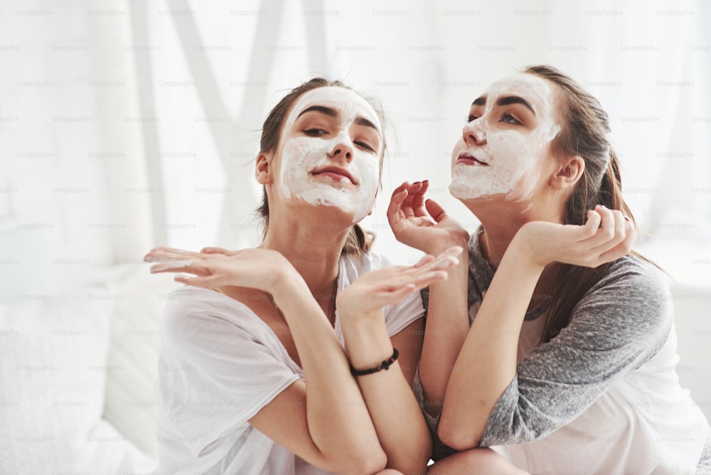 Schau uns an, wir sind großartig. Konzeption der Hautpflege mit weißer Maske im Gesicht. Zwei Schwestern haben Wochenende im Schlafzimmer.