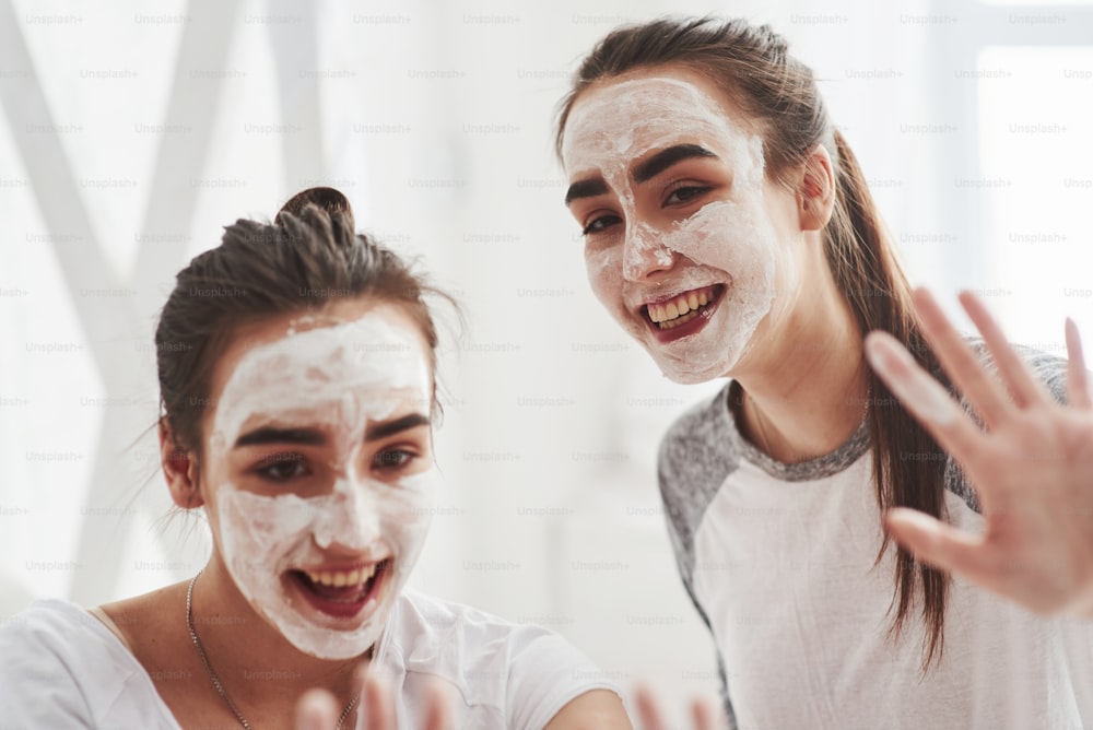 Say hello to the cameramen. Conception of skin care by using white mask on the face. Two female sisters have weekend at bedroom