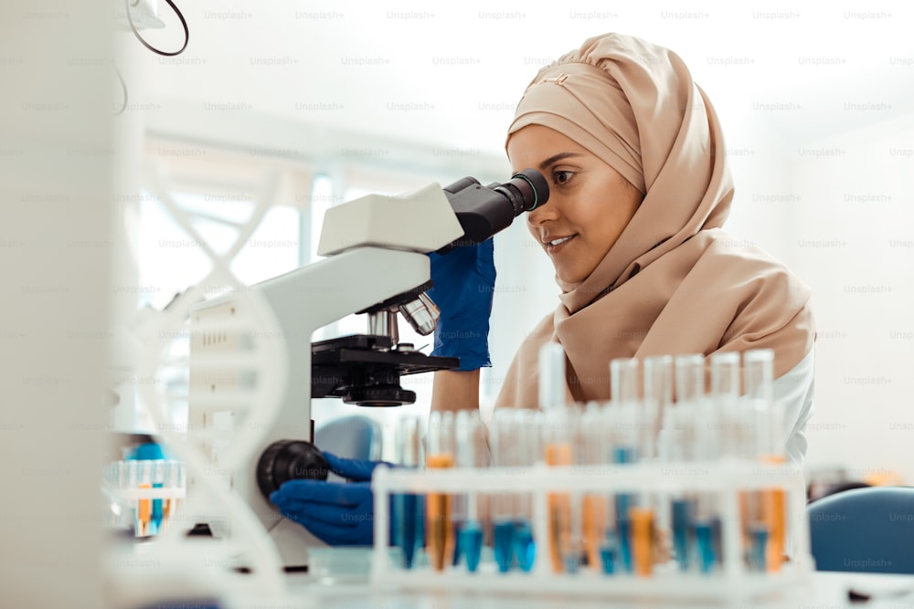 Trabajador de laboratorio. Mujer musulmana positiva mirando en el microscopio mientras está trabajando en el laboratorio