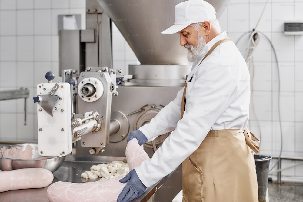 Macellaio che lavora con la produzione di salsicce, in piedi vicino all'attrezzatura, tenendo salsiccia. Lavoratore che indossa uniforme bianca, grembiule marrone, guanti di gomma. Industria alimentare.