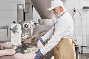 Butcher man working with production of sausages, standing near equipment, holding sausage. Worker wearing in white uniform, brown apron, rubber gloves. Food industry.