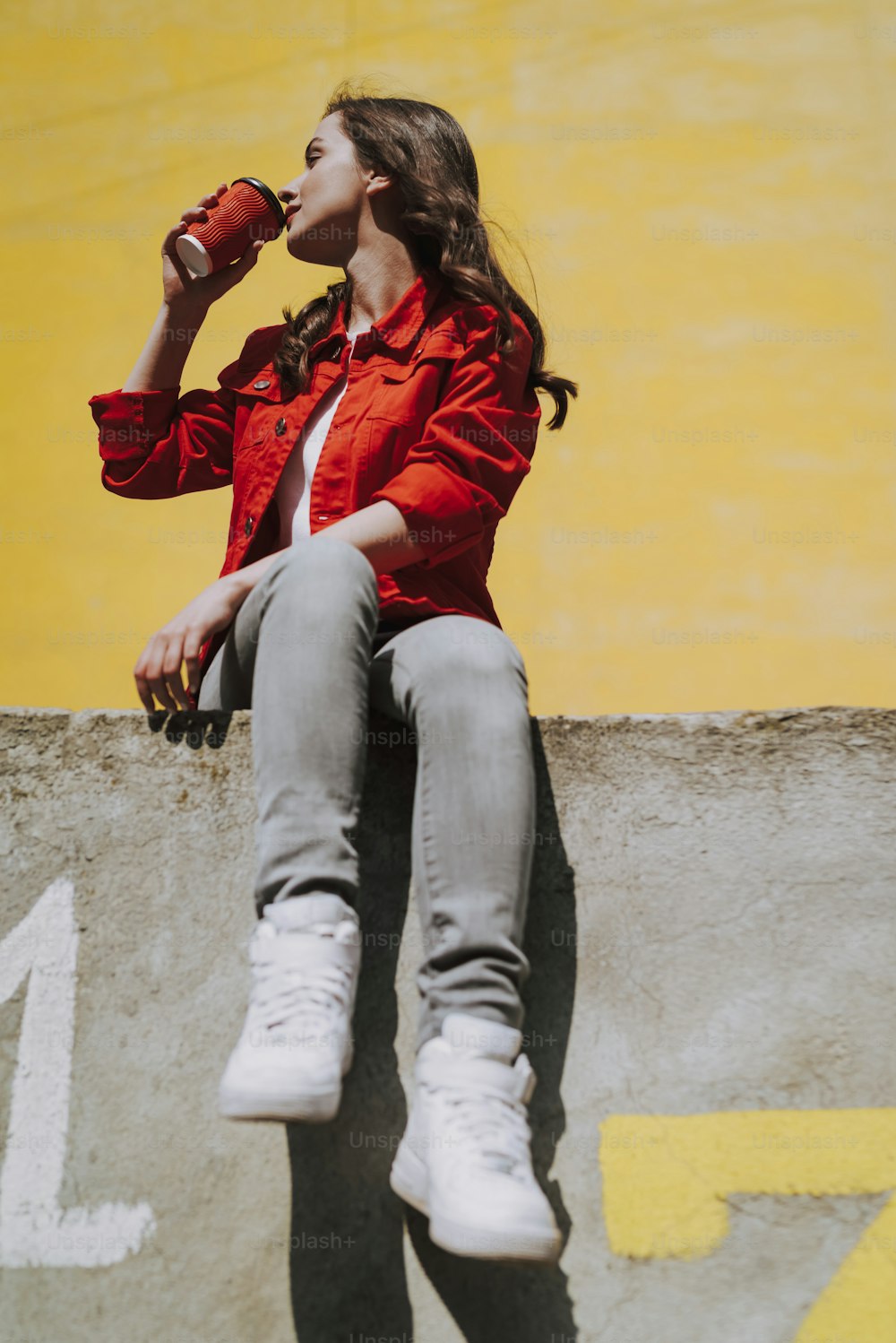 都会のライフスタイルのコンセプト。欄干の壁に座りながらコーヒーを飲む若いかなりブルネットのヒップスターの女性の低角度のフルレングスの肖像画
