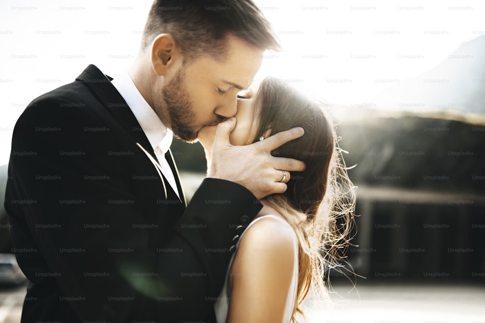 Retrato de perto de um belo jovem casal se beijando no dia do casamento contra o nascer do sol, enquanto o noivo está segurando o rosto de sua noiva.