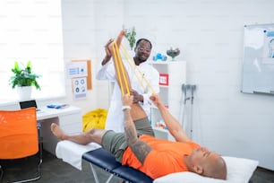 Ribbon and stretching. Bald sportsman holding yellow ribbon while stretching leg visiting therapist