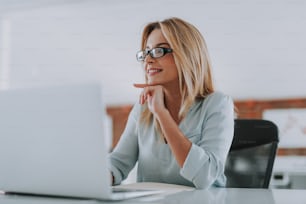 Glad office worker. Positive young woman feeling good and smiling while sitting in front of the laptop at her workplace and touching her face with one hand