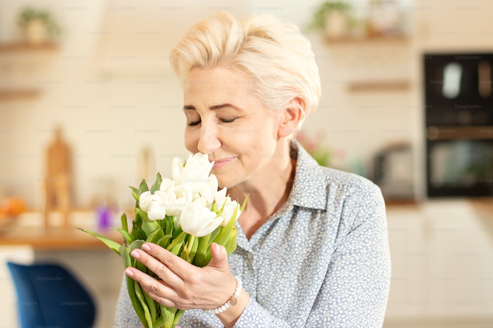 Middle age woman holding bouquet of tulips in home with a delicate smile on her face. Blonde lady with short hairstyle.