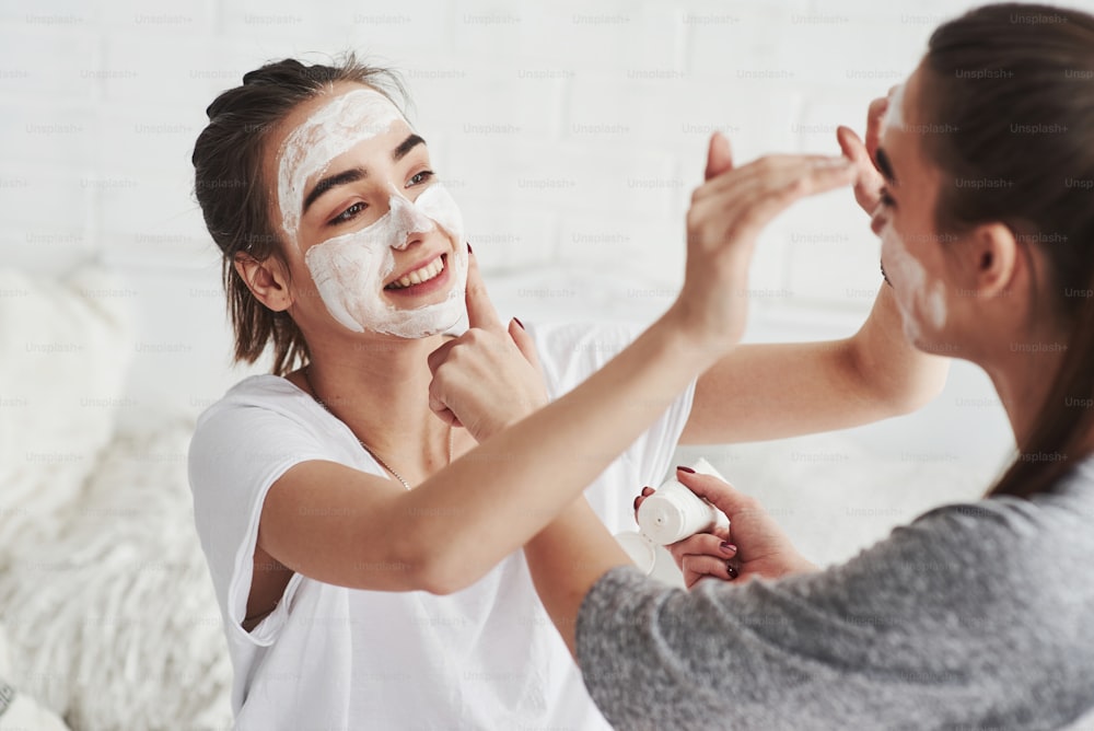 You look funny. Conception of skin care by using white mask on the face. Two female sisters have weekend at bedroom.