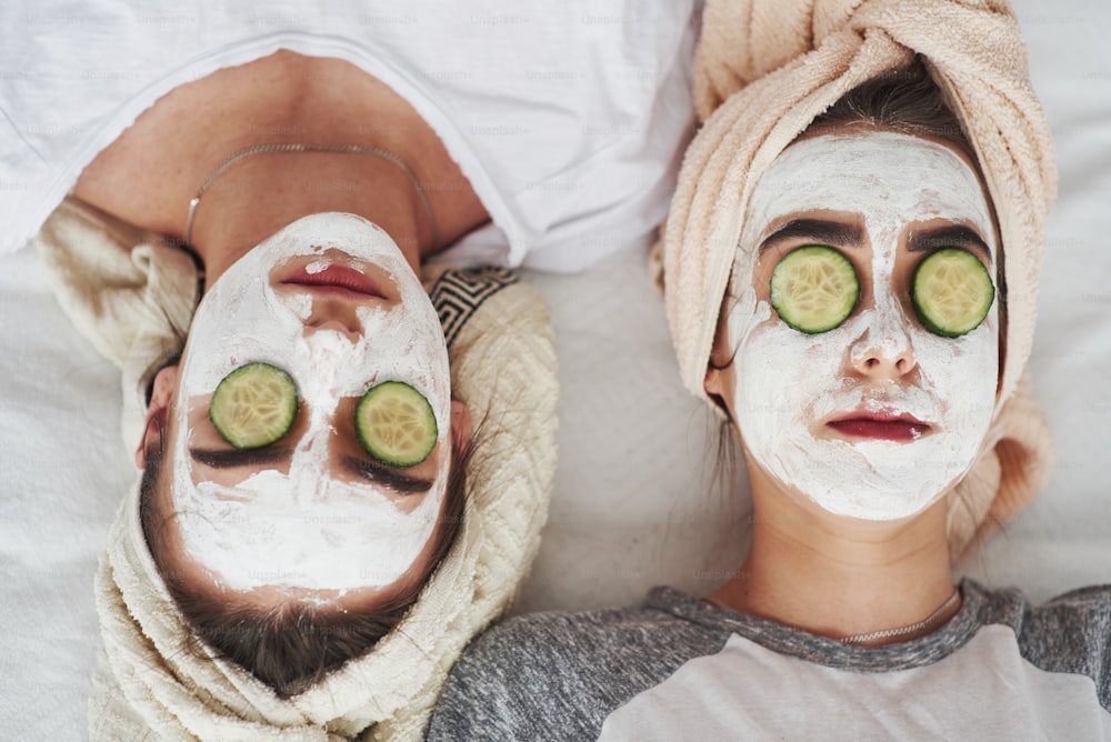 Kopfüber liegend. Konzeption der Hautpflege mit frischen Gurkenringen und weißer Maske im Gesicht. Zwei Schwestern haben Wochenende im Schlafzimmer.