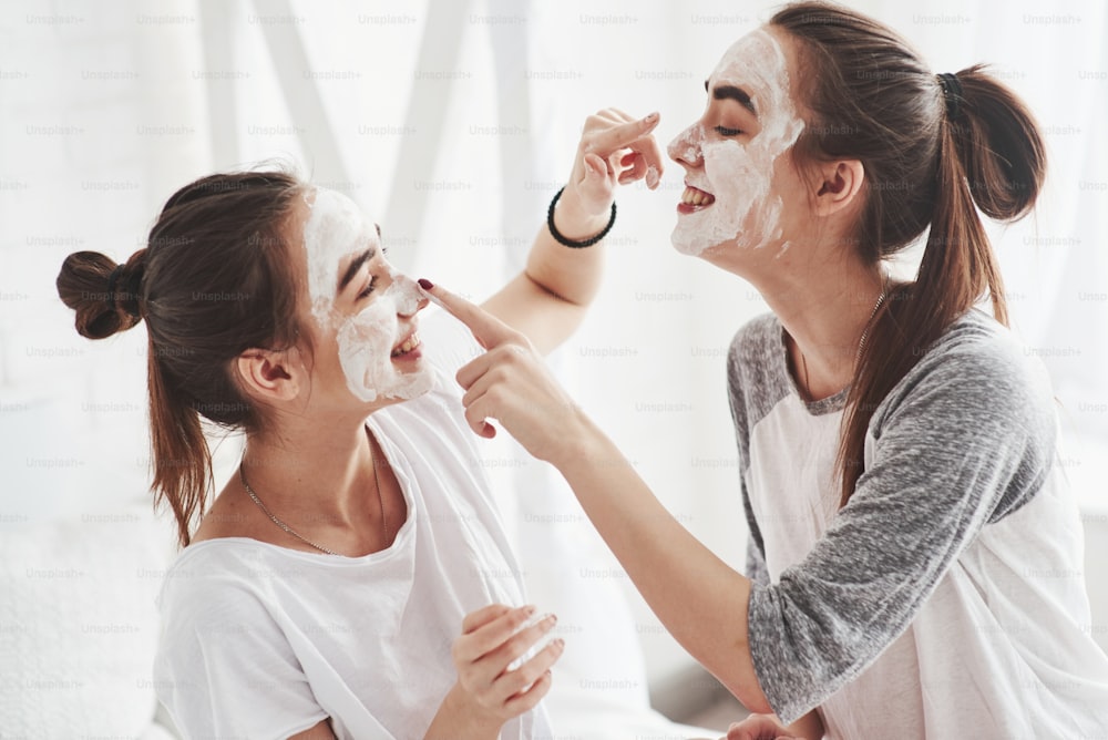 Berührung der Nase. Konzeption der Hautpflege mit weißer Maske im Gesicht. Zwei Schwestern haben Wochenende im Schlafzimmer.