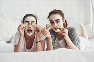 Is that beautiful or not. It will be after. Conception of skin care by using fresh cucumber rings and white mask on the face. Two female sisters have weekend at bedroom.