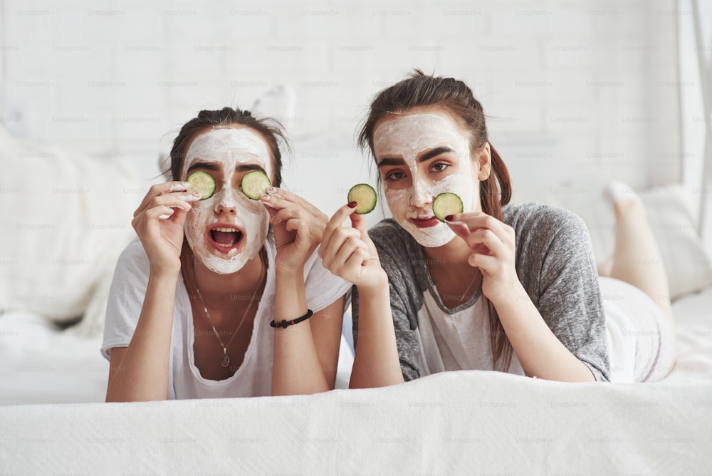Is that beautiful or not. It will be after. Conception of skin care by using fresh cucumber rings and white mask on the face. Two female sisters have weekend at bedroom.