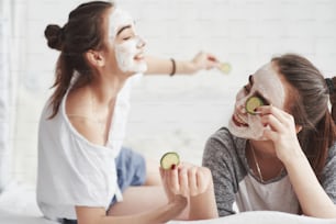 How this works. Conception of skin care by using fresh cucumber rings and white mask on the face. Two female sisters have weekend at bedroom.