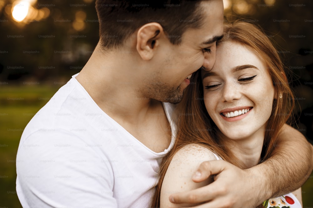 Amazing close up portrait of a cheerful caucasian red haired woman with freckles laughing with closed eyes while embracing with her man against sunset outside.