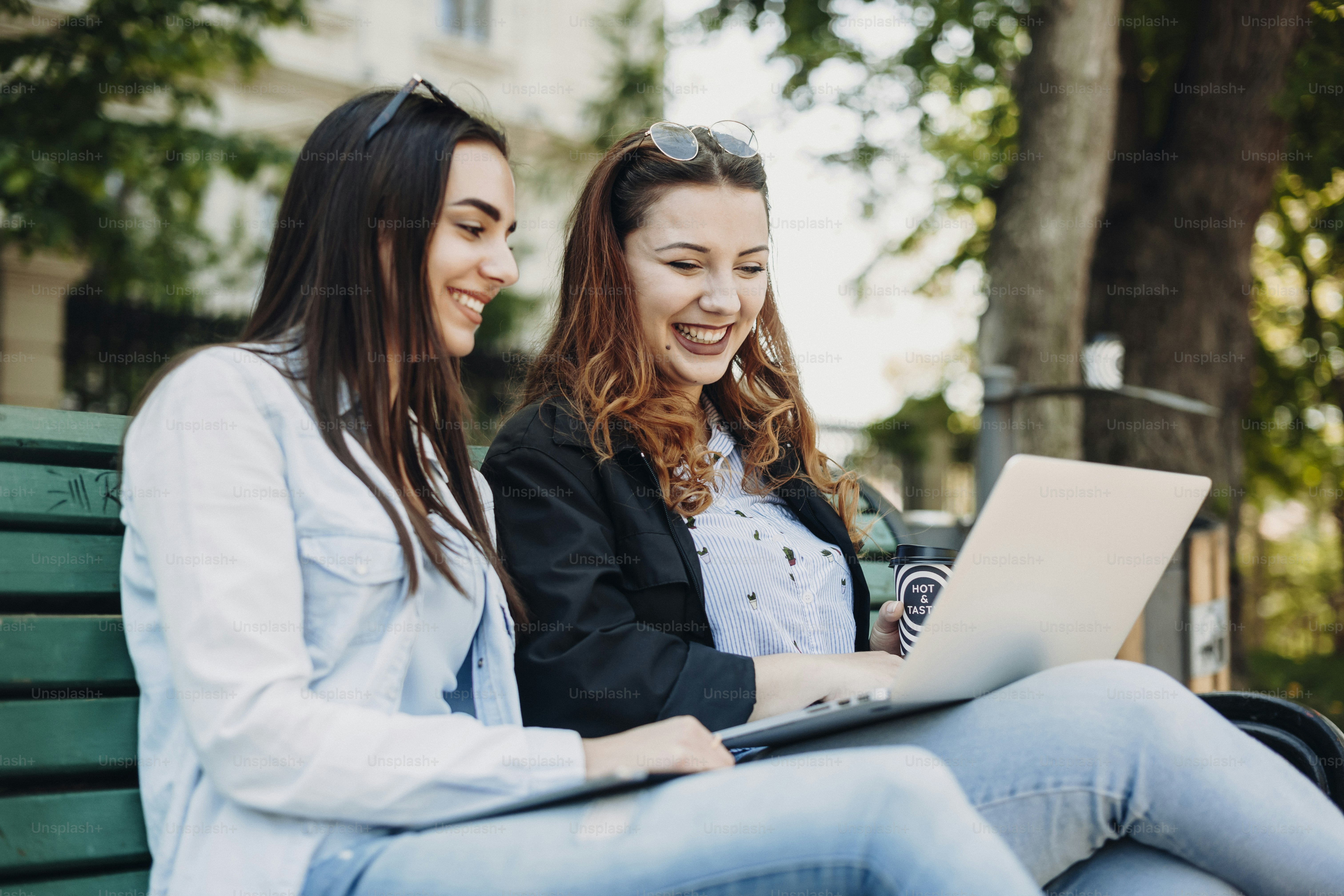 Two beautiful caucasian female sitting on a bench outside laughing while looking at a laptop holded on the legs.