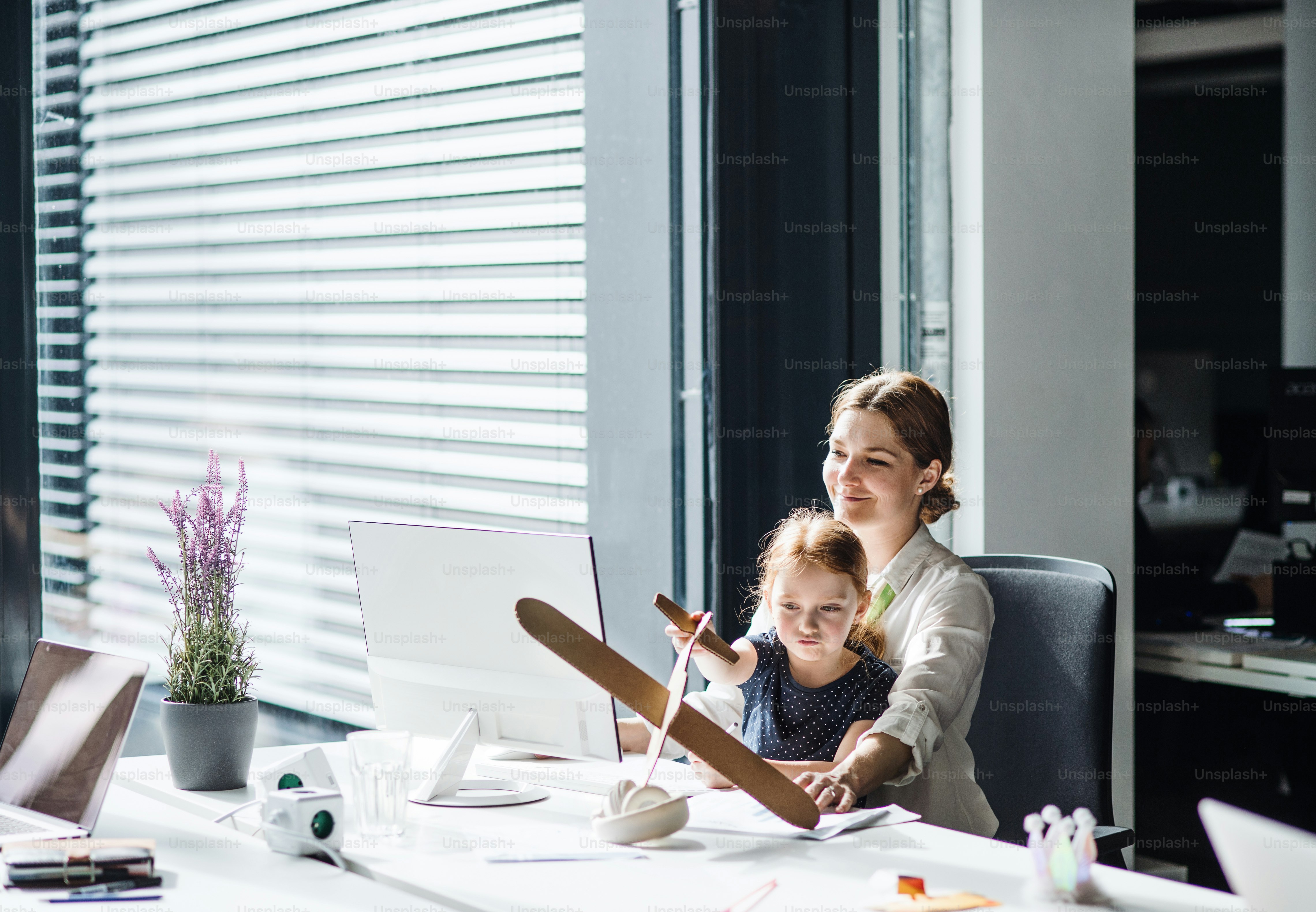 A businesswoman sitting and working in an office, a small daughter playing with a paper plane.