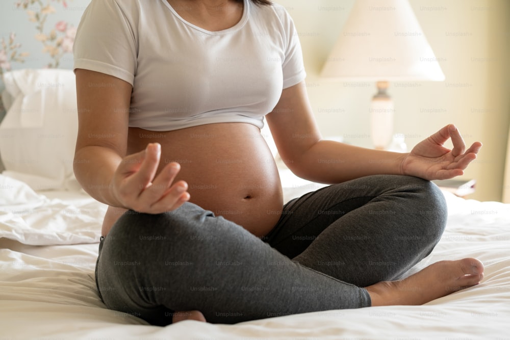 Schwangere Frau, die Yoga-Übungen auf dem Bett im Schlafzimmer zu Hause macht, während sie sich um ihr Kind kümmert. Die glückliche junge werdende Mutter, die das Baby im schwangeren Bauch hält. Schwangerschaftsvorsorge und Schwangerschaft bei Mutterschaft.