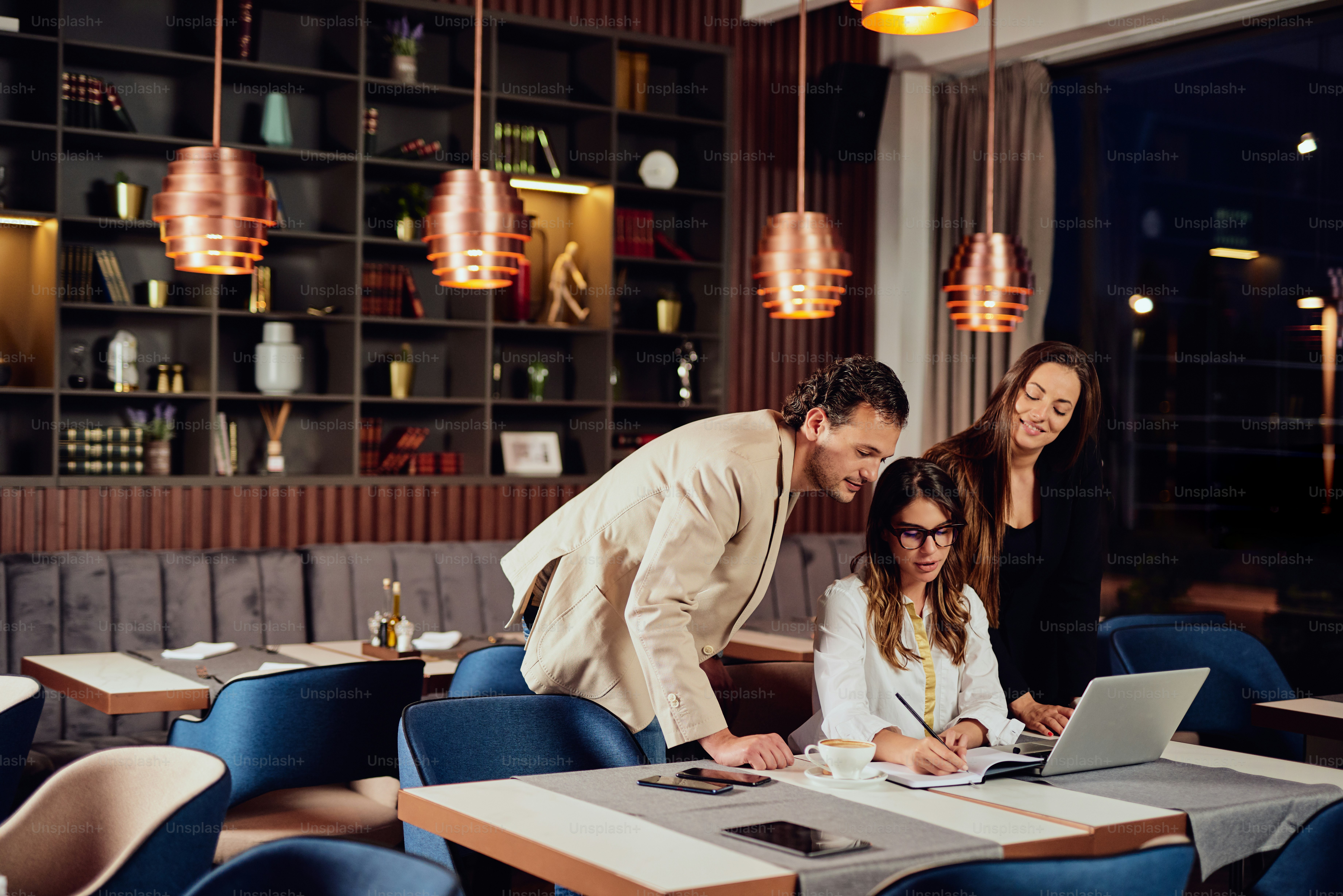 Charming buisnesswoman with brown hair and dressed elegant sitting at restaurant and looking at laptop. Next to her standing two colleagues and looking at laptop, too.