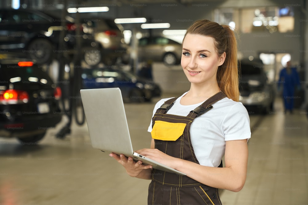 Beautiful young woman working as mechanic, holding notebook in hands. Pretty girl looking at camera, smiling, posing. Female worker wearing in white t shirt and brown coveralls.
