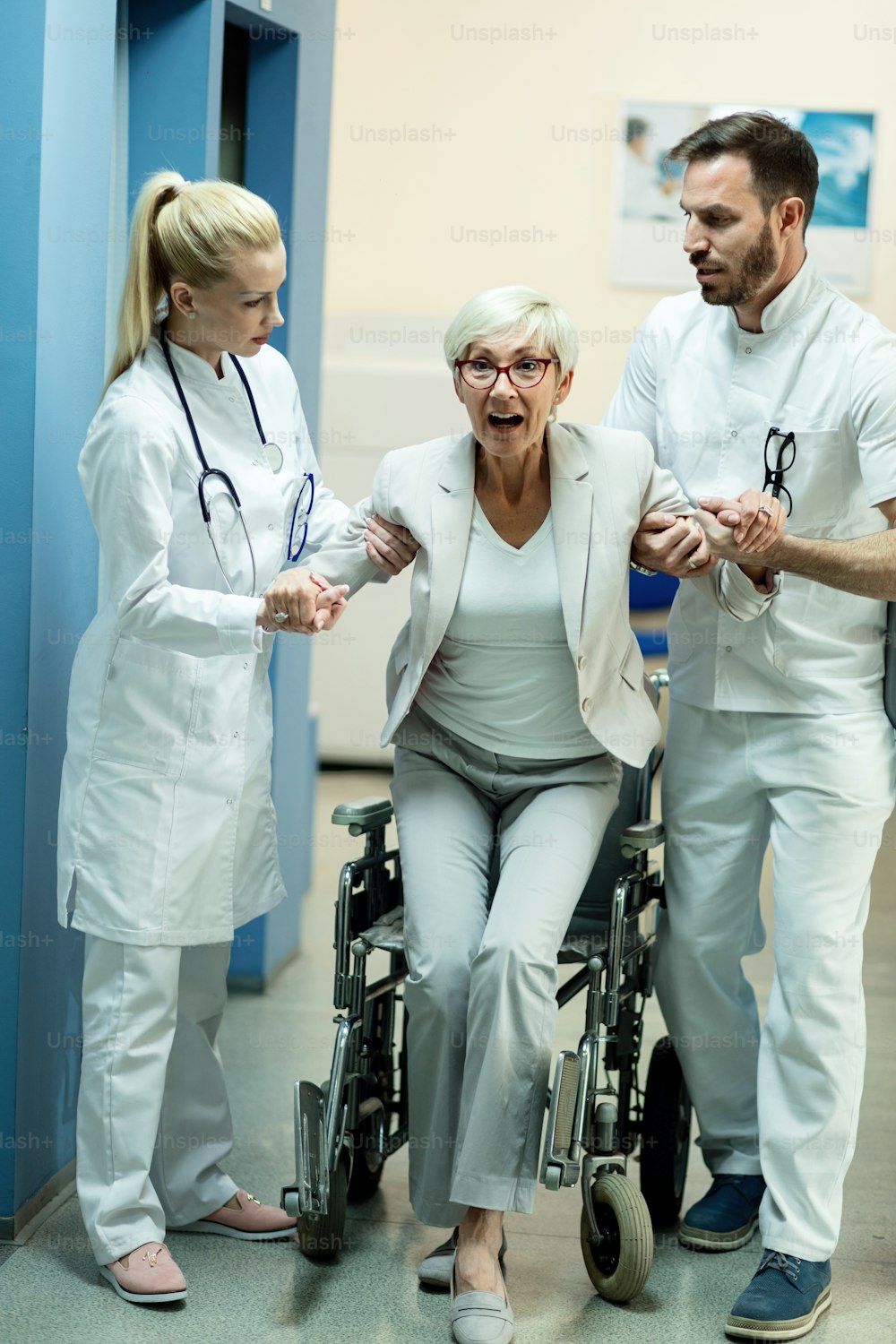 病院内の2人の医師の助けを借りて車椅子から立ち上がり、驚いた表情を浮かべる障害のある女性患者。