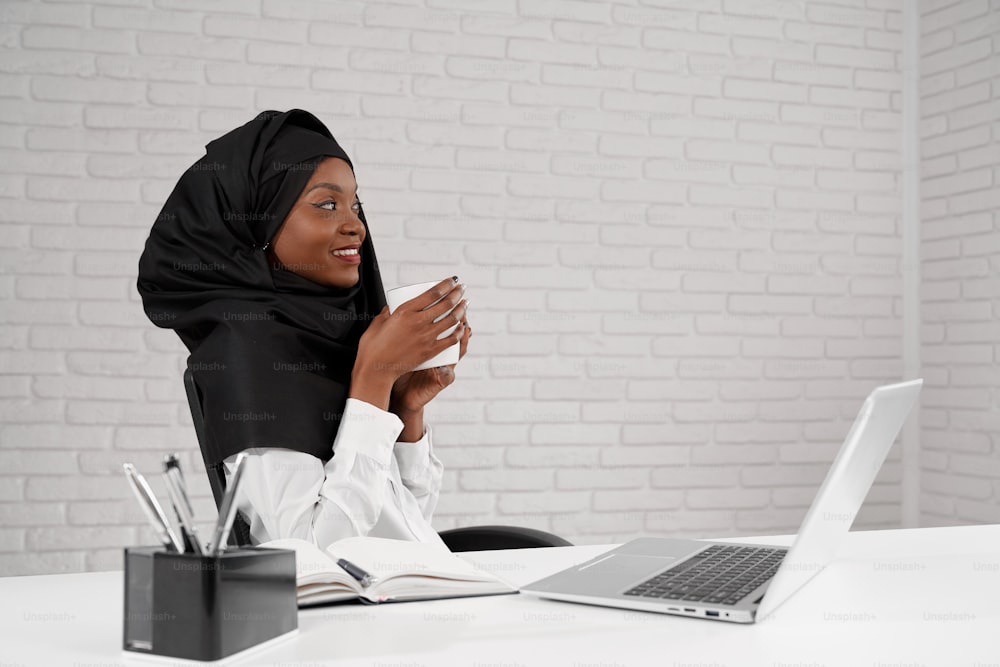Side view of smiling woman wearing black hijab drinking coffee and looking at window while having break at job. Beautiful african lady working in multi ethnic company. Concept culture and success.