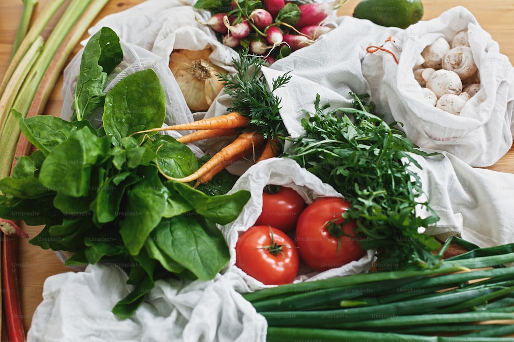 Wiederverwendbare umweltfreundliche Taschen mit frischem Gemüse Karotten, Tomaten, Spinat, Rucola, Pilze, Rhabarber, Zwiebeln auf Holztisch. Plastik verbieten. Zero-Waste-Einkaufskonzept für Lebensmittel