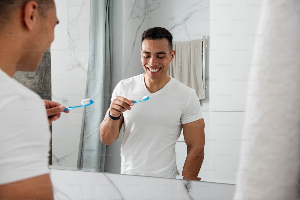 Morning hygiene concept. Back side portrait of young muscular happy male in white t-shirt looking at tooth brush while staying in bathroom before mirror