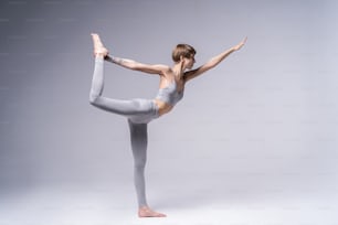 Portrait of attractive happy young woman working out indoors, doing yoga exercise, variation of Natarajasana, Lord of the Dance, King Dancer or Standing Mermaid Pose