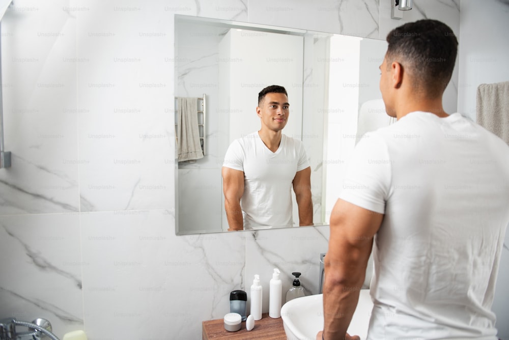Morning hygiene concept. Back side waist up portrait of young brunette muscular smiling guy in white t-shirt looking at his reflection on mirror in bathroom