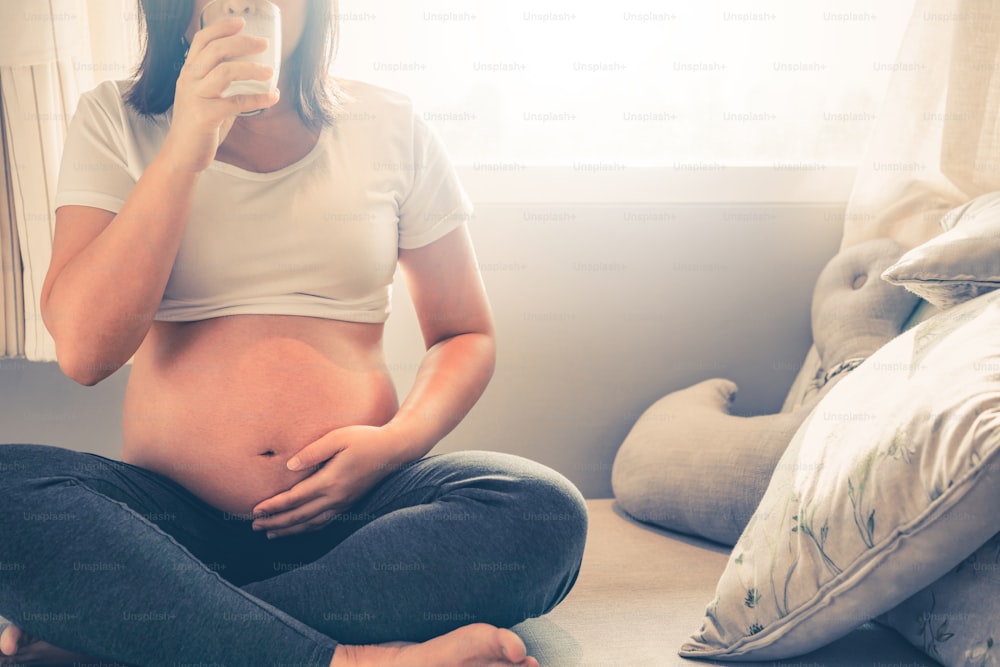 Donna incinta felice beve latte in vetro a casa mentre si prende cura di suo figlio. La giovane madre in attesa che tiene il bambino nella pancia incinta. Nutrimento alimentare a base di calcio per ossa forti della gravidanza.