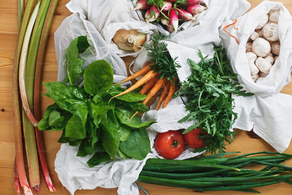 Zero waste grocery shopping concept,flat lay. Reusable eco friendly bags with fresh vegetables carrots,tomatoes, spinach,arugula, mushrooms,rhubarb,onions on wooden table. ban plastic.
