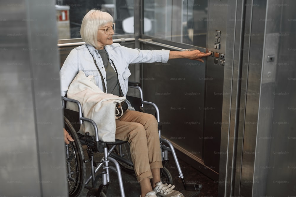 Full length portrait of old female holding white bag and sitting in wheelchair while pushing elevator button
