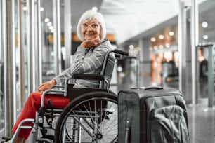 Foto recortada de mujer adulta sentada en silla de ruedas en el aeropuerto con computadora portátil y maleta y esperando registrarse mientras sonríe