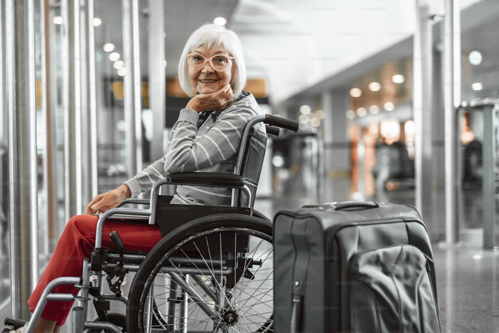 Cropped photo of adult woman sitting in wheelchair at airport with laptop and suitcase and waiting check in while smiling