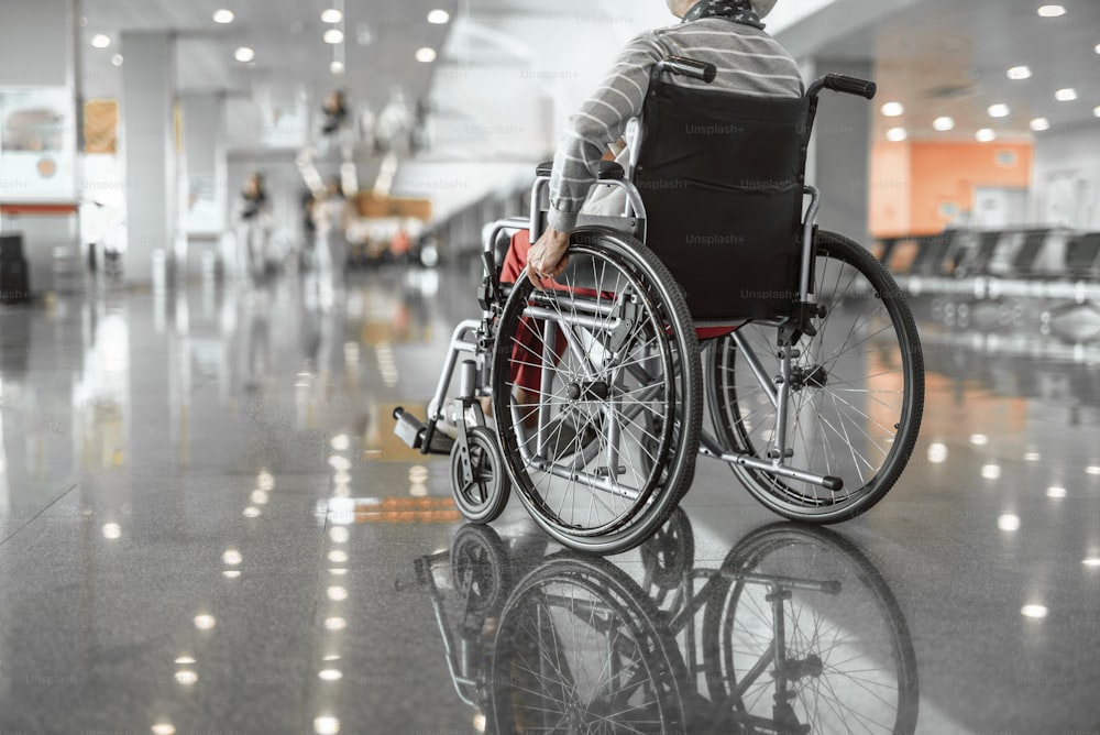Back view photo of old woman moving in wheelchair in the lobby at airport. Copy space in left side