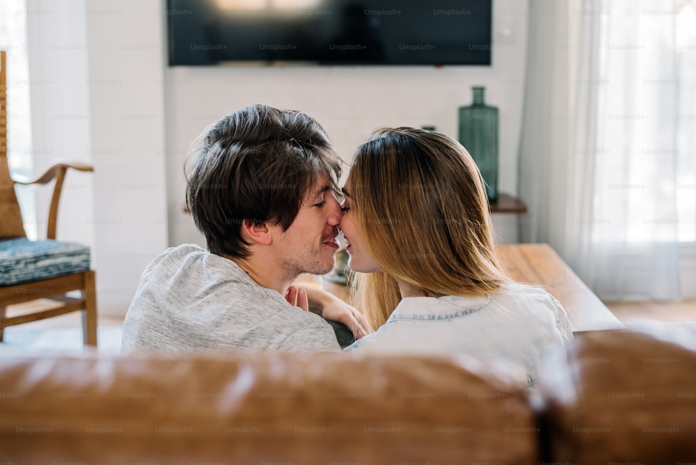 Encantadora pareja joven abrazándose besándose y mirándose en el cómodo sofá de su casa
