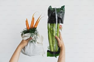 Choisissez un produit sans plastique. Mains tenant un sac écologique réutilisable avec des carottes fraîches contre du céleri dans un emballage en plastique cellophane sur fond blanc. Épicerie zéro déchet. Bannissez le plastique.