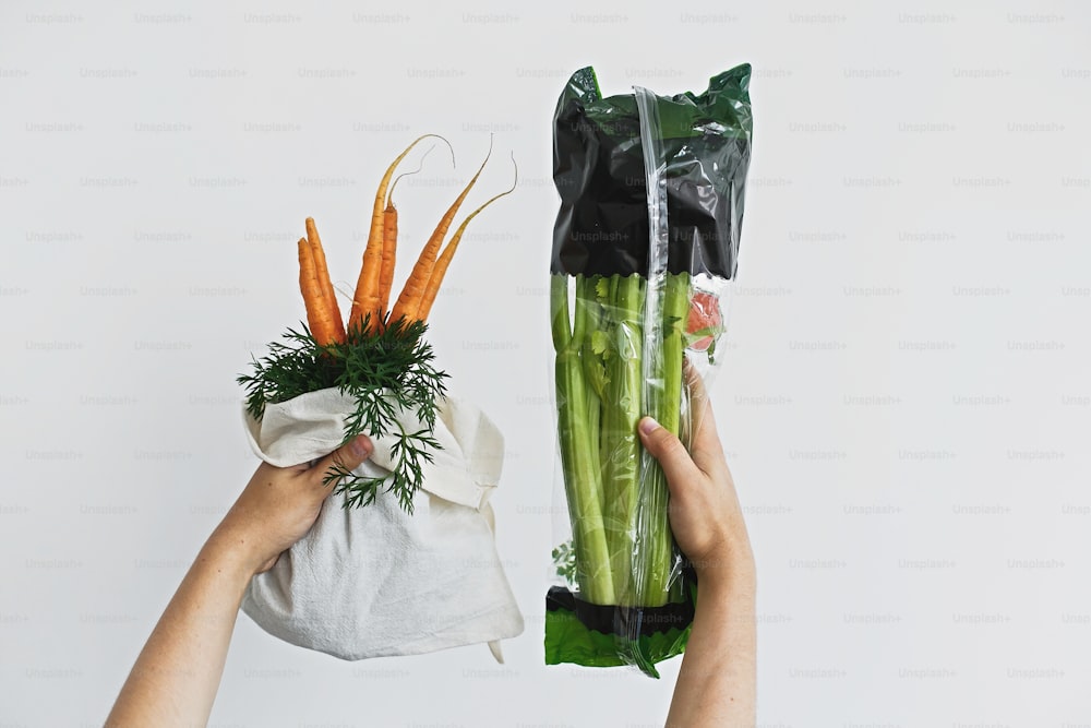 Choose plastic free. Hands holding reusable eco friendly bag with fresh carrots against celery in cellophane plastic package on white background. Zero waste grocery shopping. Ban plastic.