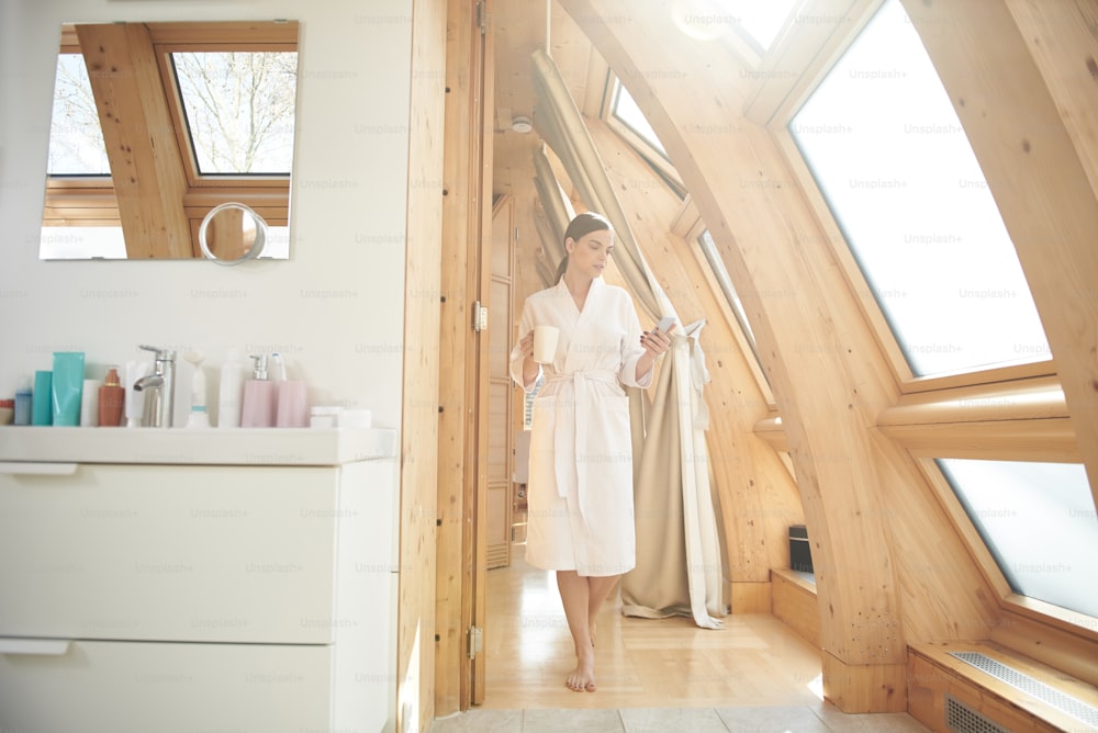 Full length of thoughtful young woman in bathrobe holding cup of tea and looking at screen of smartphone while walking barefooted in her own house