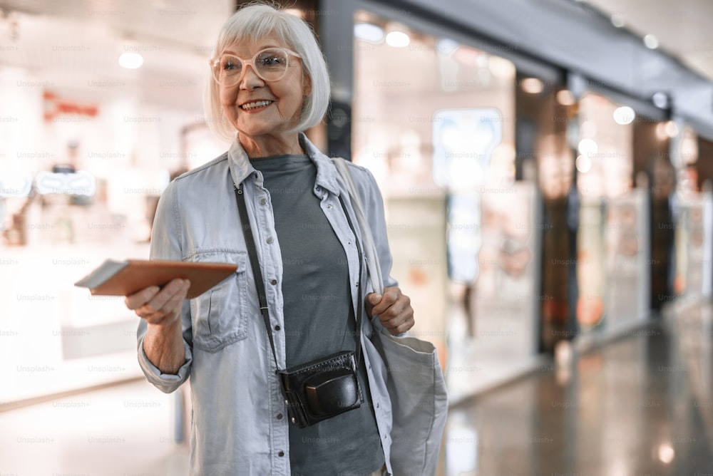 Waist up portrait of happy old woman with camera standing at airport while holding tickets. Copy space on right side