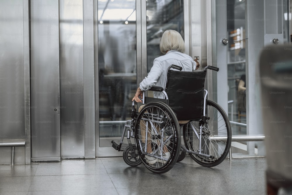 Back view portrait of old female sitting in wheelchair while and waiting for the elevator. Copy space in left side