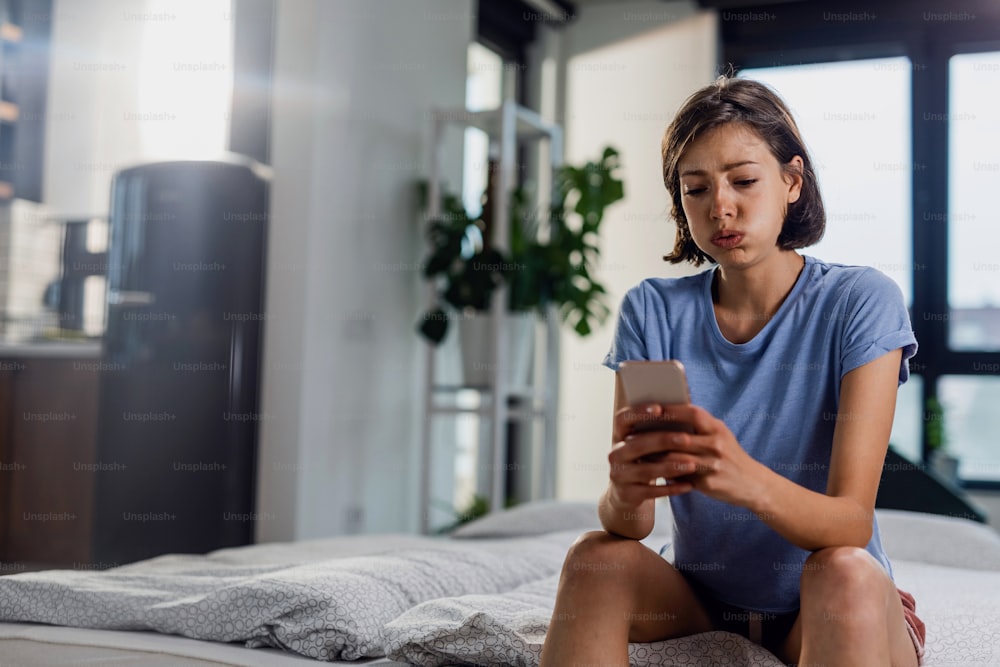 Young sad woman reading bad news on mobile phone while sitting alone in bedroom.